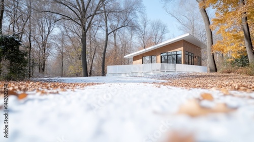 Modern minimalist house with a slanted roof stands in a snowy forest surrounded by trees with autumn leaves.