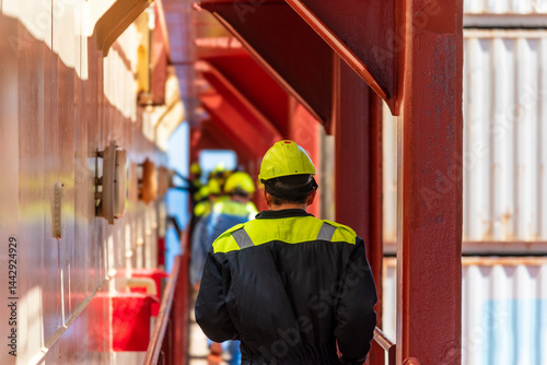 Deck officer seen from behind walking between containers and superstructure during an abandon-ship drill, dressed in blue coveralls and yellow helmet - real action on a working merchant ship.