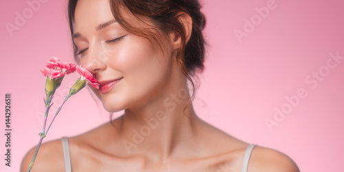 Soft and romantic portrait of a young woman gently smiling with her eyes closed while enjoying the fragrance of pink carnations. The pastel pink background, natural glow, and serene expression.