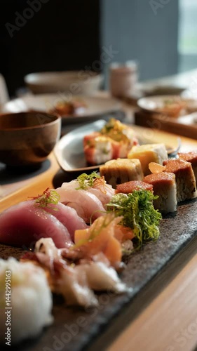 Assorted sushi nigiri presented on a slate plate, featuring tuna, squid, and various toppings served with wooden bowl and tableware.