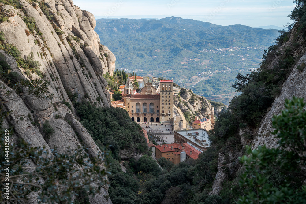 Obraz premium Views of the Monastery of Montserrat, Barcelona, Spain, from the Santa Maria gorge, surrounded by rugged rocky cliffs and dense vegetation, with distant mountains under a clear blue sky