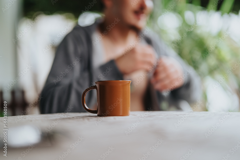 A brown mug sits on a wooden table with a blurred young male figure in the background, evoking a serene outdoor atmosphere. A brown mug sits on a wooden table with a blurred young male figure in the background, evoking a serene outdoor atmosphere.