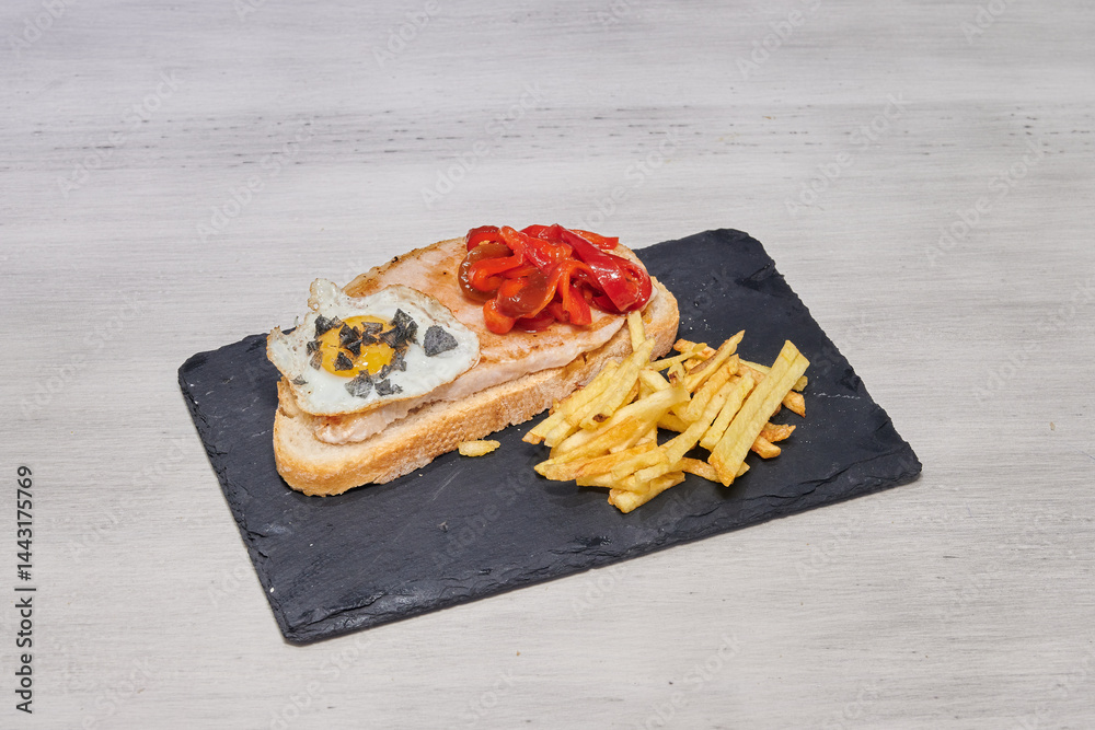 A delicious meal consisting of a sandwich, french fries, and an egg is served on a black plate. The sandwich is made with bread, ham, and cheese, while the fries are crispy and golden brown
