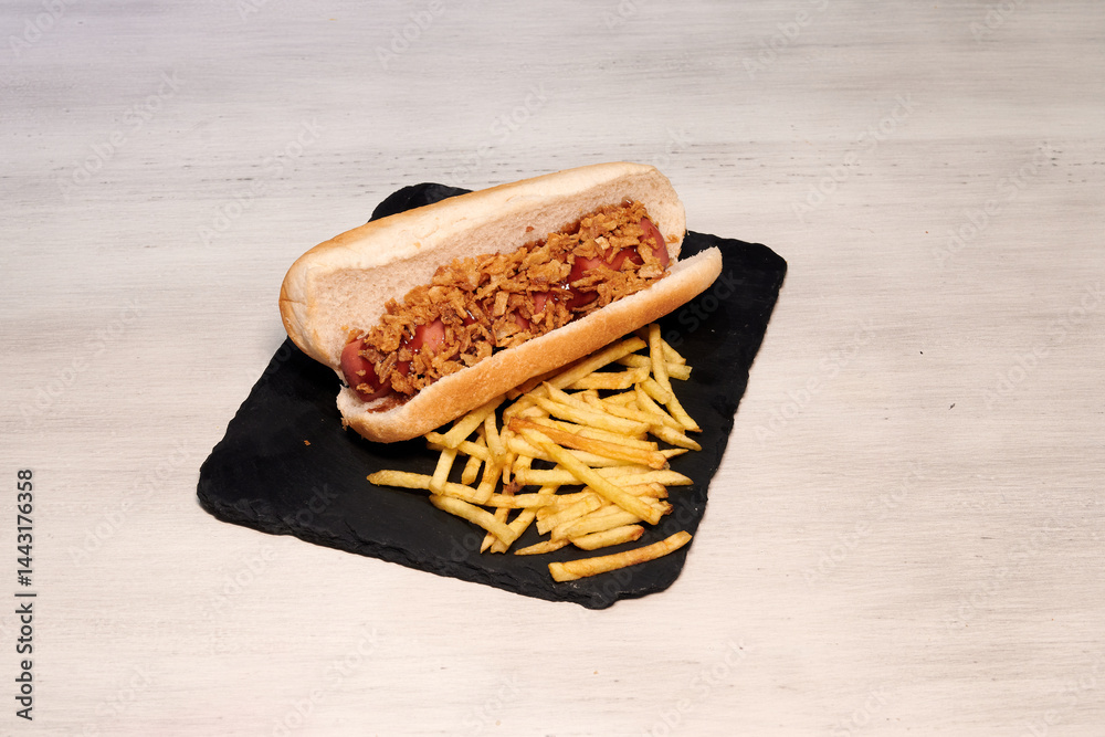 A hot dog with bacon and onions is on a black plate. There are also french fries on the plate
