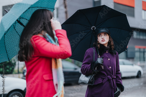 Two fashionable women wearing coats and holding umbrellas interact on a rainy day in a modern city setting, showcasing urban life and friendship in inclement weather.