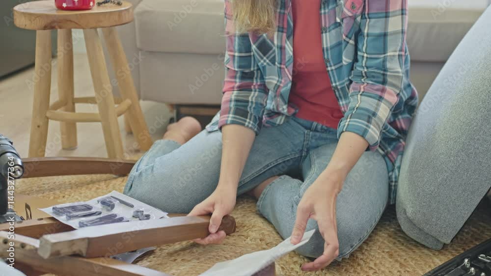 Woman sitting on floor and reading instruction manual while assembling wooden furniture