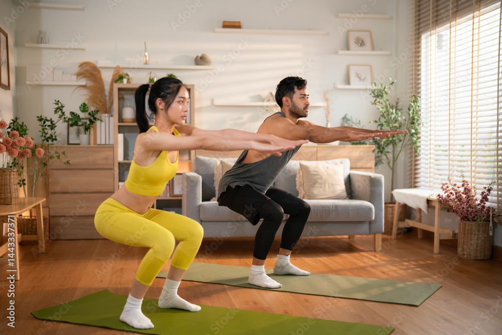Young active couple performing squats side by side in bright home setting, engaged in synchronized fitness routine. Young active couple performing squats side by side in bright home setting, engaged in synchronized fitness routine.