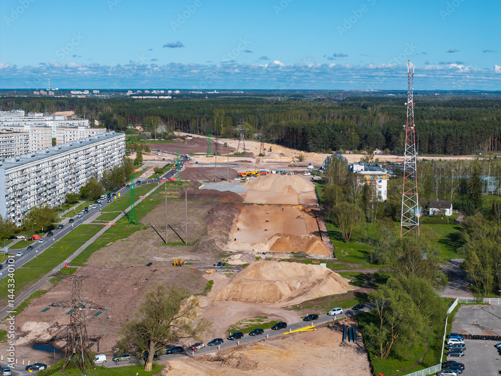 Construction site in Riga with sand mounds, equipment, a residential building, a metal tower, forested background, and urban structures in the distance.
