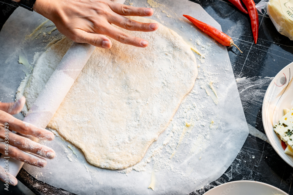 Making homemade pizza with three types of cheese - women's hands in action. Close-up scene of women's hands rolling out homemade pizza dough on a kitchen countertop.