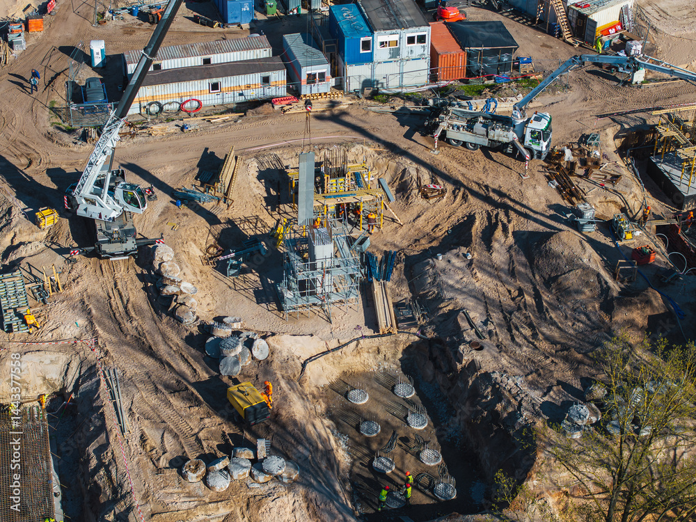 Active construction site with cranes, scaffolding, concrete piles, and workers in high visibility clothing in Riga, Latvia. Portable buildings in the background.
