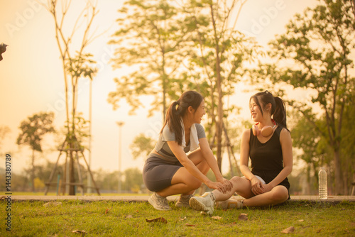 Two young Asian women jogging on a street in a park in the evening under the beautiful golden sunlight, their legs hurt so they sat down to rest. Healthy exercise concept.