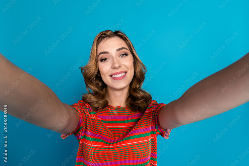 Fototapeta premium Smiling young woman with curly hair wears colorful striped shirt and poses happily against a vibrant blue background in cheerful mood