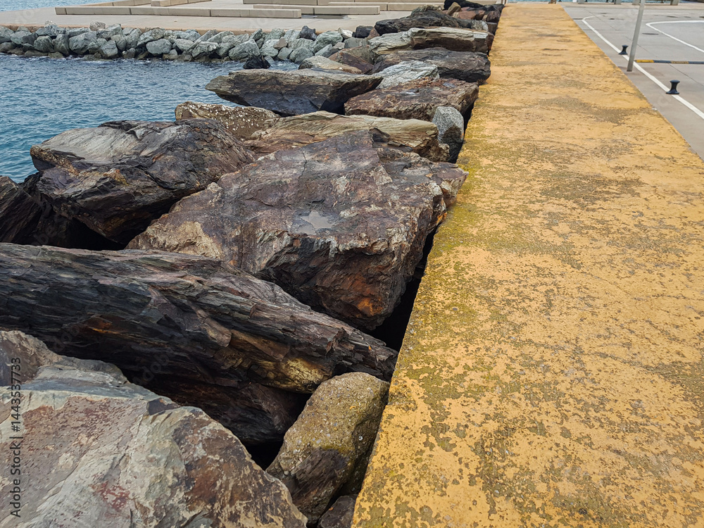 Large rocks line the edge of a waterfront pathway, with a yellow-painted surface and calm water in the background, creating a tranquil coastal atmosphere