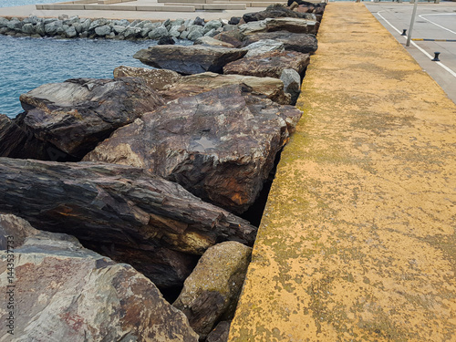 Obraz na płótnie Large rocks line the edge of a waterfront pathway, with a yellow-painted surface and calm water in the background, creating a tranquil coastal atmosphere w salonie