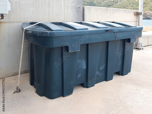 A robust green waste container is fastened with a strap, positioned beside a concrete wall near a scenic coastal location.