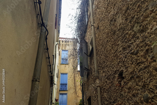 Narrow alleyway displays old buildings with blue shutters and textured stone walls. Soft daylight filters through, illuminating the architectural details of the historic town