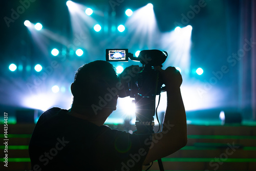 Silhouette of a cameraman operating a video camera on a tripod, capturing a live event under bright blue stage lighting with lens flare and glowing bokeh in the background.