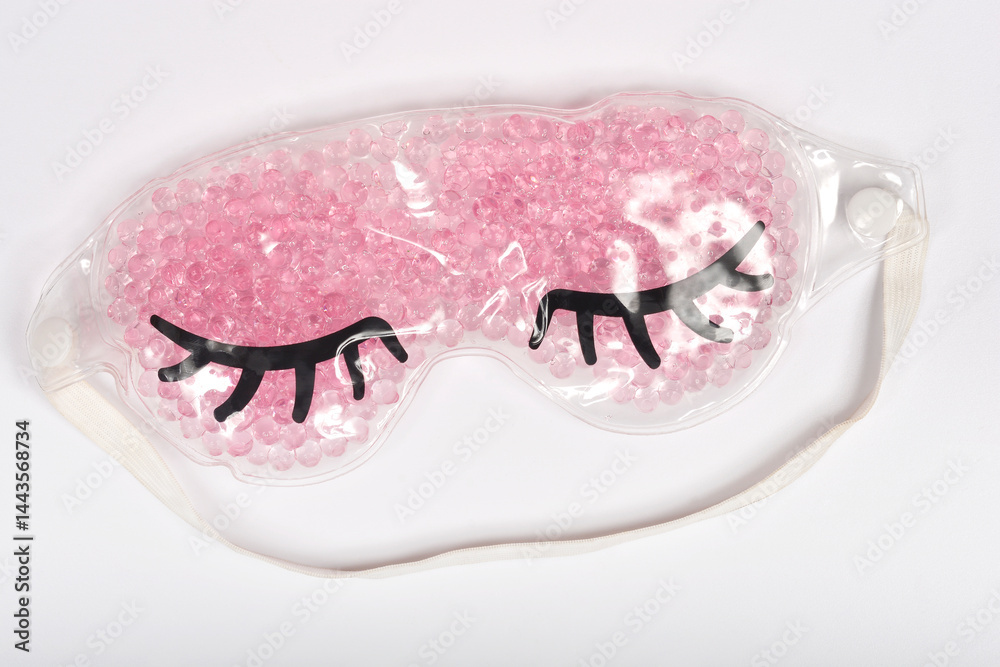 A clear gel eye mask filled with small pink beads and featuring cartoon black eyelashes rests on a light gray surface