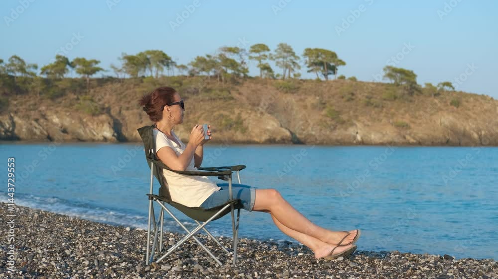 Woman relaxing on beach, sipping from cup and enjoying view. Gentle waves lap at the shore as a woman sits in a folding chair on a pebble beach, savoring a drink and taking in the serene view Woman relaxing on beach, sipping from cup and enjoying view. Gentle waves lap at the shore as a woman sits in a folding chair on a pebble beach, savoring a drink and taking in the serene view