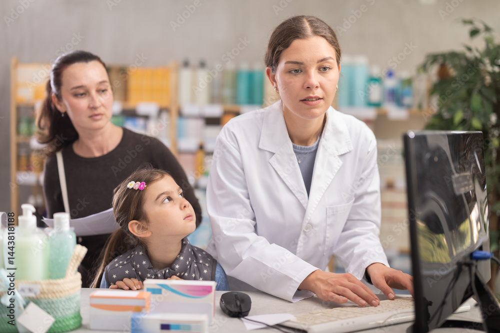 Fototapeta premium Child and mother with doctors prescription visiting drug store. At cash register area with PC, girl pharmacist consulting clients, prepare customer using manual.