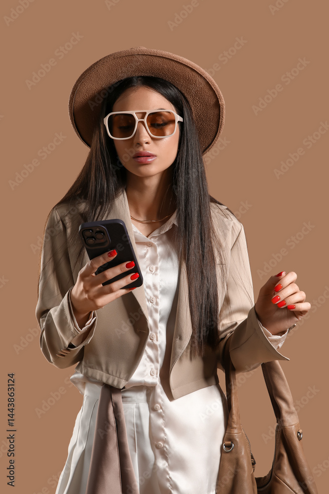 Fototapeta premium Stylish young woman in sunglasses using mobile phone on brown background