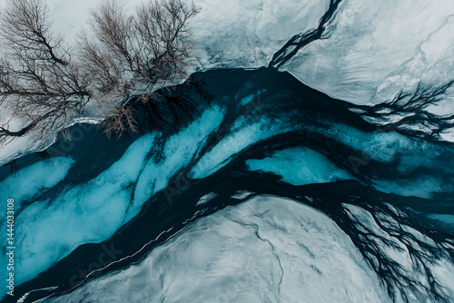 A minimalist aerial photo of the landscape, showing a dark sandbank and an icy river flowing through a snow-covered environment.