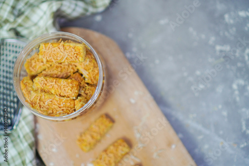 top view of an open jar filled with cheese cookies and some cookies lined up outside the jar, with cheese grater, gingham fabric and wooden chopping board below it