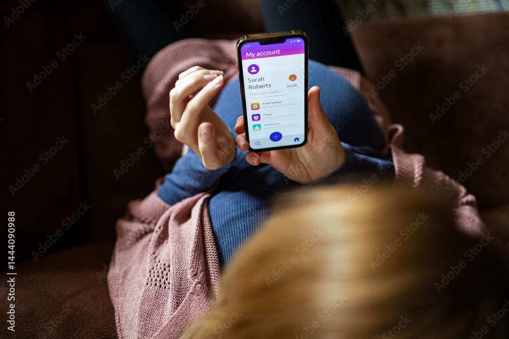 Pregnant woman using mobile banking app on smartphone while relaxing on couch