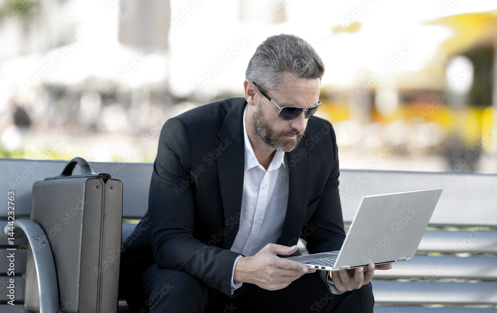 Confident business man sitting on a bench with a laptop and phone in park. Successful business man. Mature businessman enjoying work break. Businessman taking a break on a park bench. Summer business.