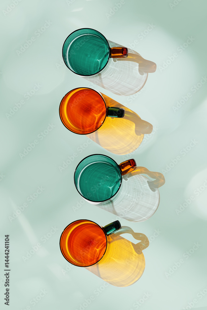 Orange and green glowing glass cups on teal background, artistic light reflections and shadow in natural sunlight. Summer drink concept, minimal trend pattern of transparent glasses, caustics