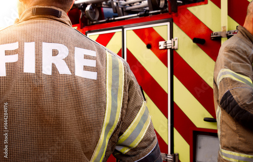 Firefighter team - firemen equipped with emergency services response firefighting crew uniform and gear near fire engine. Department red fire truck appliance from fire station is in scene background.