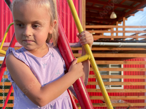 Little girl on playground holds colorful bars near modern wooden house.
