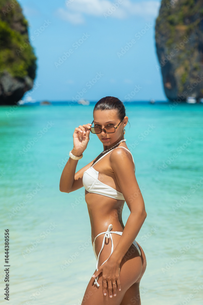 Stylish woman influencer with glowing skin and slim figure posing confidently in white bikini with sunglasses at Maya Bay, Thailand, concept of fashion, summer travel, lifestyle and high quality promo