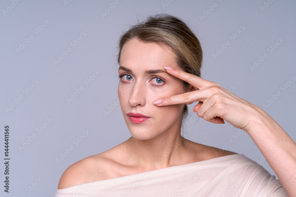 Beautiful young woman in pastel clothes makes a gesture around her eyes, emphasizing the freshness of her skin, the beauty of her look and her facial care