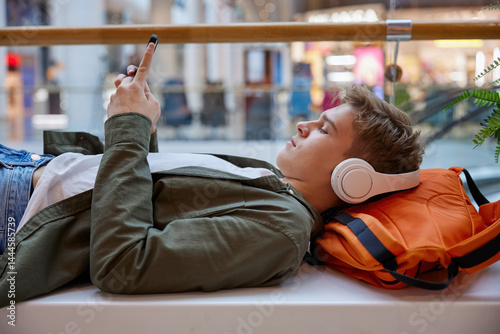 Obraz na płótnie Teenage boy listening to music using smartphone resting in shopping mall w salonie