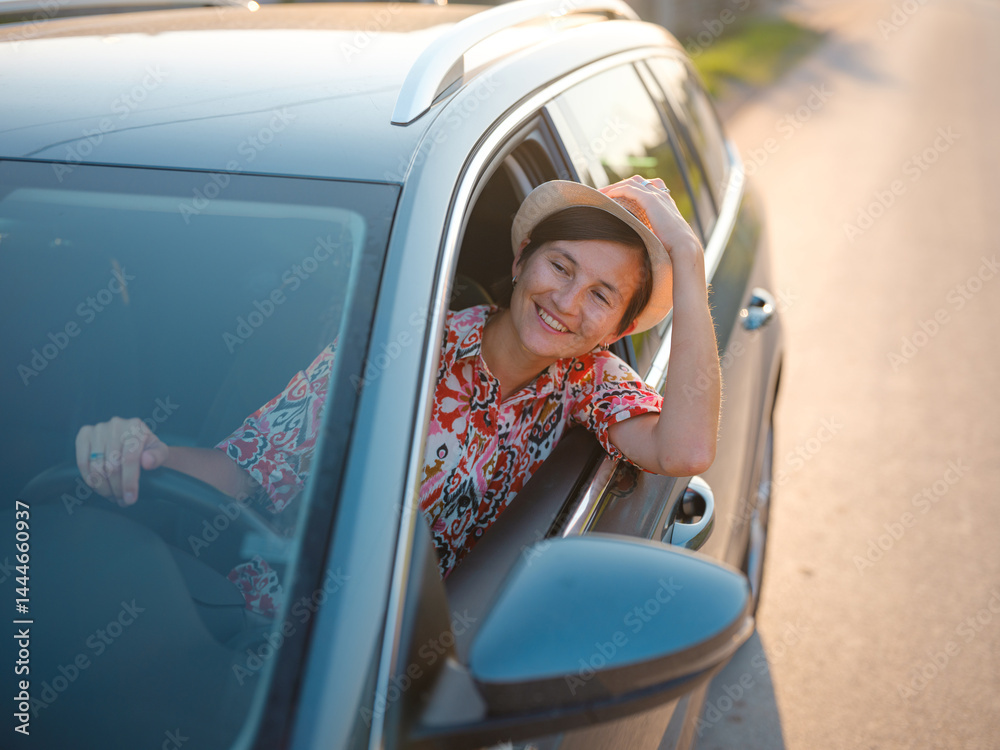 Naklejka premium Traveler woman in red ethnic dress and hat drives car through scenic countryside of Croatia in summer. Surrounded by lush fields and forests, she enjoys peaceful and adventurous road trip