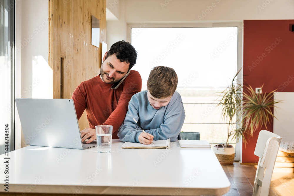 Father multitasking with laptop while son writes in notebook at home Father multitasking with laptop while son writes in notebook at home
