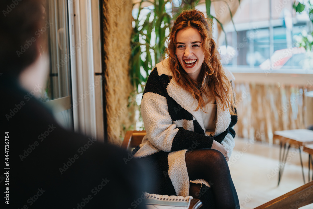 Cheerful young woman sitting inside a cafe with natural light and a happy expression. A warm environment enhances the pleasant interaction during a relaxed and positive atmosphere.