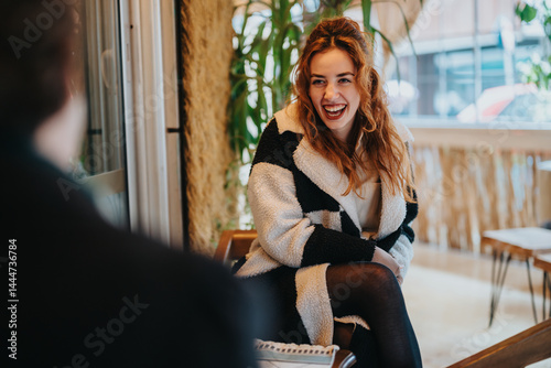 Obraz na płótnie Cheerful young woman sitting inside a cafe with natural light and a happy expression. A warm environment enhances the pleasant interaction during a relaxed and positive atmosphere. w salonie