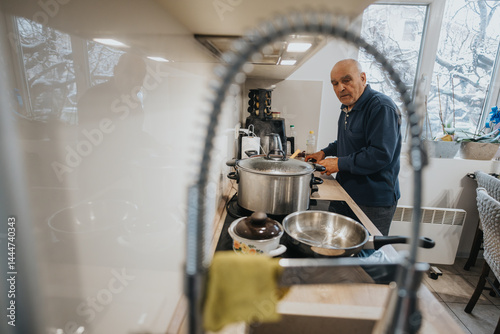 Obraz na płótnie An elderly man preparing food in a modern kitchen, creating a homely atmosphere. Snow visible through the windows emphasizes the warm and cozy indoor setting focused on culinary activity. w salonie