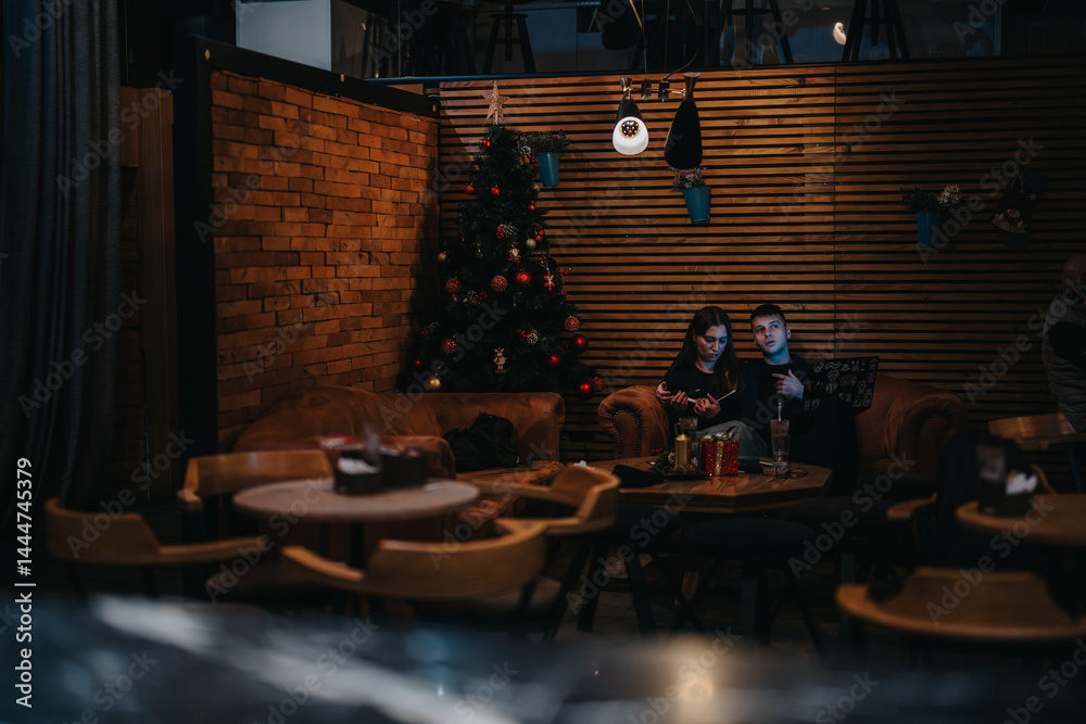 Two individuals relax in a warmly lit cafe, sitting on a sofa near a beautifully decorated Christmas tree, creating an intimate and festive setting for sharing moments or working together.