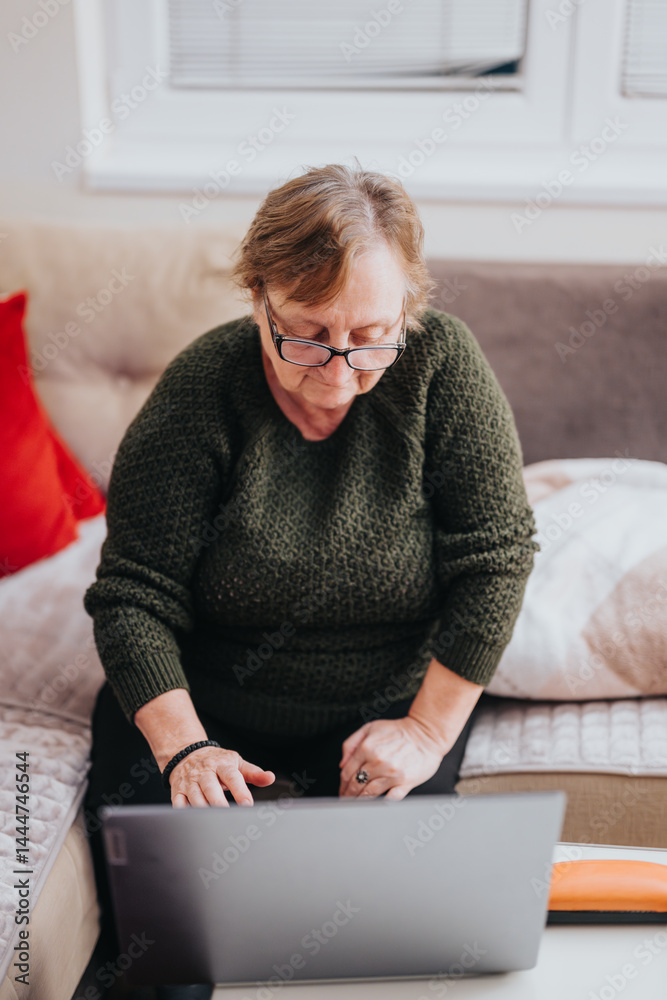 An elderly woman wearing a green sweater works on her laptop while sitting on a modern couch in a warmly lit interior.