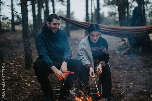 A pair of friends enjoy roasting sausages near a campfire in a winter forest, surrounded by a tranquil ambiance and nature's beauty.
