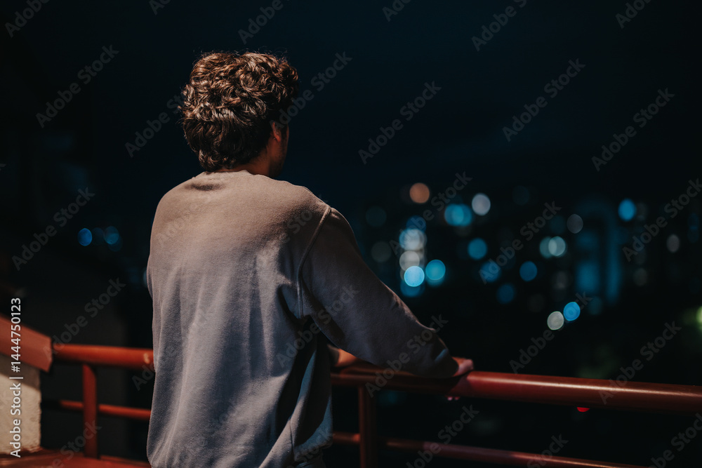 A young man stands on a balcony, gazing thoughtfully at the glowing city lights against the night sky, conveying a sense of reflection and solitude.