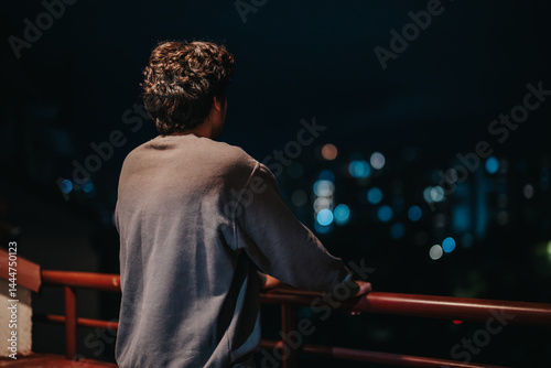 Obraz na płótnie A young man stands on a balcony, gazing thoughtfully at the glowing city lights against the night sky, conveying a sense of reflection and solitude. w salonie