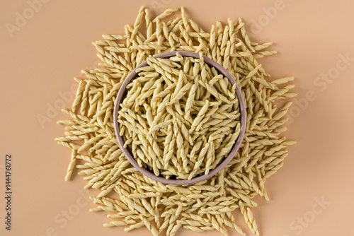 Top view of a bowl of raw italian durum wheat trofie pasta against a beige background .