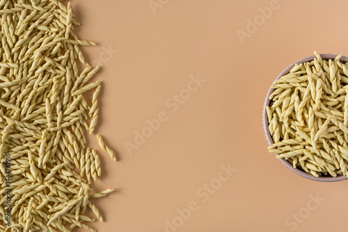 Top view of a bowl of raw italian durum wheat trofie pasta against a beige background .