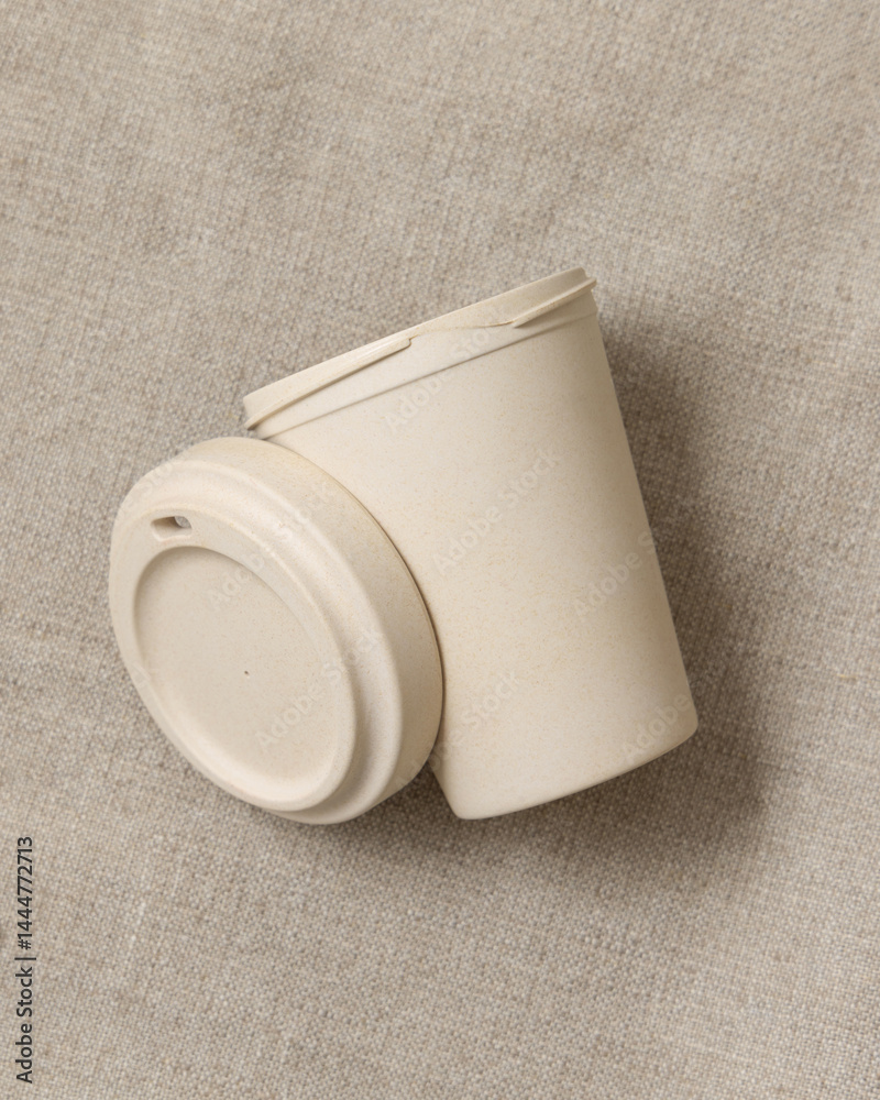 Opened beige reusable bamboo coffee cup on beige linen cloth top view, copy space, mockup