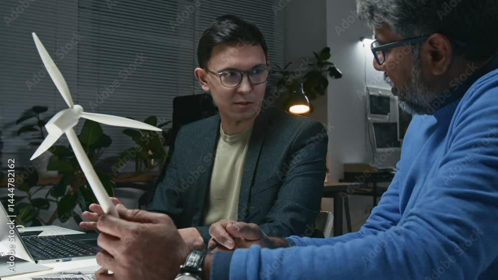 Medium shot of Asian businessman talking to mature engineer with wind turbine model in hand, discussing setup of sustainable technologies on site Medium shot of Asian businessman talking to mature engineer with wind turbine model in hand, discussing setup of sustainable technologies on site