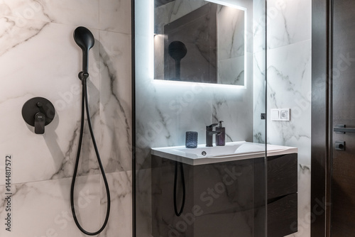 modern bathroom design in luxury apartments. walls are decorated with marble tiles in shades of gray and white. large mirror, sink, brown cabinet under sink, black faucets. shower cabin 